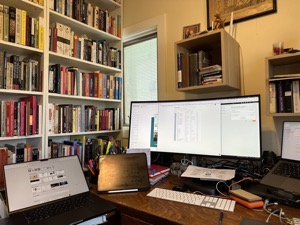 A view of my desk showing the floor to ceiling bookcases to the side, two laptops, a big wide monitor and an iPad. Over the desk are a couple Ikea shelves with assorted dictionaries and some decorative items and moderate clutter scattered about.