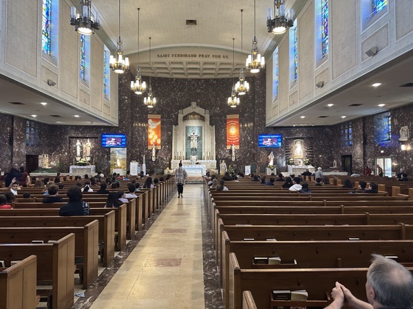 View of the interior of St Ferdinand church from the back The altar is visible straight ahead along with pews that are filled with spaced children from the parish school. The church itself is a rectangular floorplan, typical of mid-century Catholic church architecture.