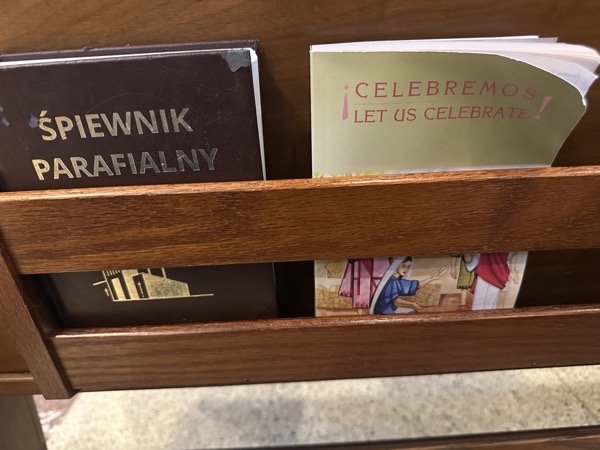 Two worship aids in the back of a pew. On the left, is a hardcover with a title of Śpiewnik Parafialny (Polish for “Parish songbook”). On the left is a paperback with a bilingual title of “¡Celebramos/Let us celebrate!”