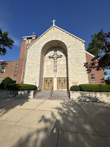 The front entrance of Sacred Heart church. There’s a tall arch over the main doors with a large cross under the arch. A bell tower is on the right and a stone cross is positioned at the peak of the roof. The facade is stone and brick.