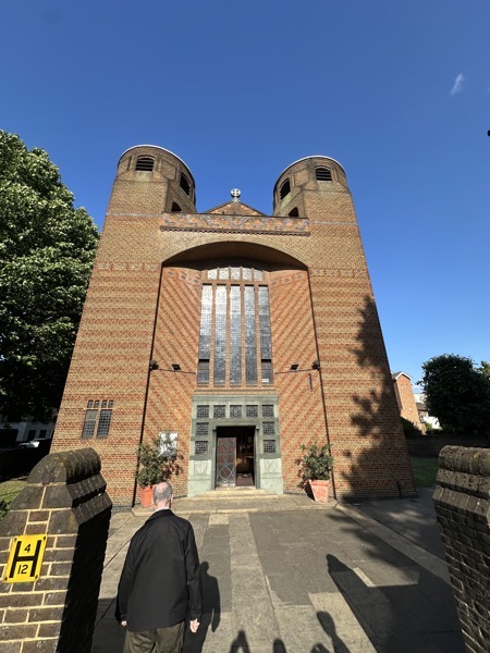 The view of the front of the church, which is a Grade II listed building. It has a facade of patterned brick with twin bell towers and a tall window over the main entrance