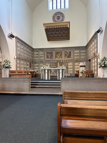 The sanctuary as visible from my pew. There is a canopy suspended over the altar and on the back wall, the Holy Trinity symbol with “Deus” at the center, with paths reading “est” leading out to “Pater”, “Filius” and “Spiritus Sanctus” that are on a circle, all joined by “non est.” Behind the altar are a triptych of pictures showing Jesus in the manger, healing a sick man (or maybe raising Lazarus?) and rising from the dead.