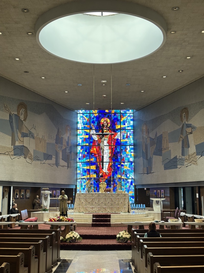 The view of the sanctuary, a raised circular platform. It’s hard to see in the image, but there is a sort of modernist altar rail surrounding the sanctuary. I would guess that the altar, under the cloth that covers it, displays the sort of curvilinear design that’s evident in the ambo and the baptismal font. On the clerestory walls over the sanctuary is a semi-abstract mosaic of a Bible scene on either side of a large stained0glass window showing the resurrected Christ.