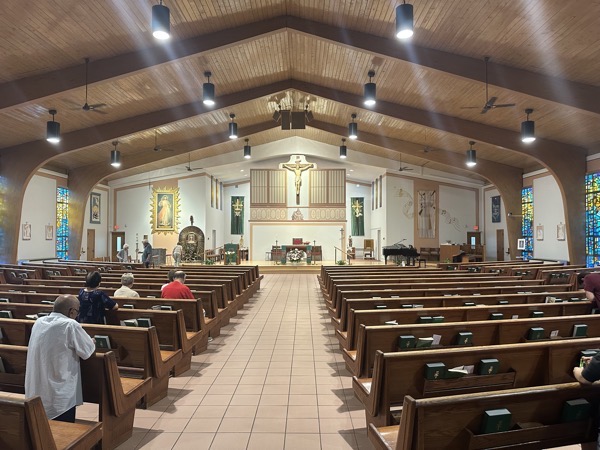 View of the sanctuary of St Colette church. The church was built in the 1950s and reflects the aesthetics of the time. The original sanctuary is now a chapel space behind a divider (an “open air” confessional is visible to the left). The choir loft is now overflow seating with the choir instead seated at the front of the church on the right.