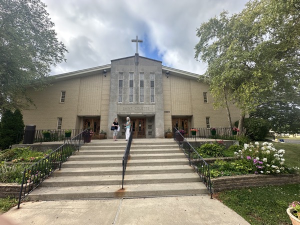The front of St Colette’s church. The façade is brick and stone with concrete steps leading up a slight hill to the entrance. There are a handful of narrow windows on an otherwise solid façade and a stone cross is mounted at the peak of the roof at the front of the church.