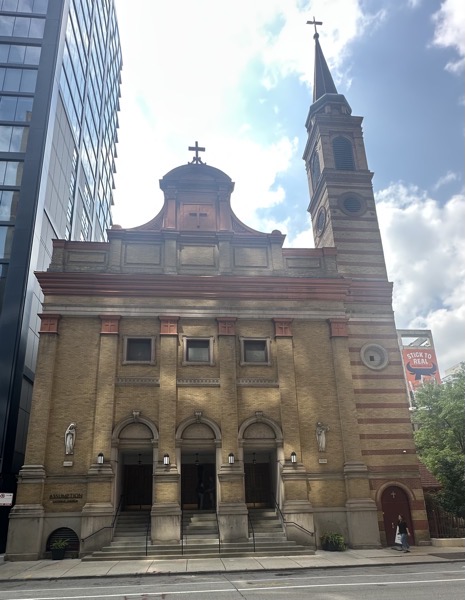 The façade of Asssumption church, which is nestled next to a glass and steel skyscraper. The church itself is a moderately sized building made of brick with a granite foundation and some stone decorative elements, Statues of Mary and Joseph adorn the front of the church/
