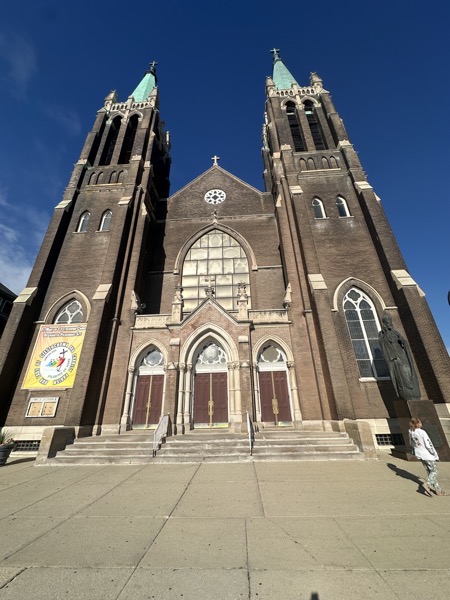 The front of St Mary of Częstochowa showing its two massive bell towers dominating the neo-Gothic façade built in brick. To the left, a sign proclaims the church a Jubilee pilgrimage site, to the right, a statue of Christ the King by Polish sculptor Czesław Dźwigaj stands.