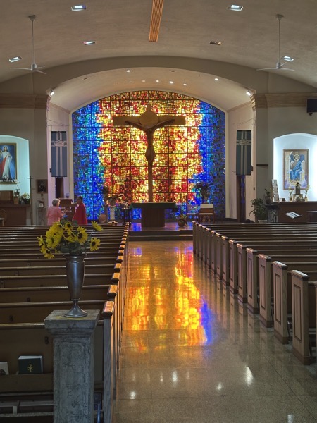 The view of the interior of the church. The back of the sanctuary is dominated by a wall composed entirely of stained glass in an abstract image which in the morning hours is lit dramatically by the sun behind it. An enormous crucifix stands against it. 