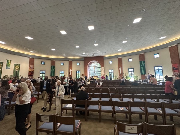 the interior of the church, The large transparent windows along the far side of the church revealing the wooded area behind the church is something I’ve never encountered before. In lieue of pews, the church has movable chairs sans kneelers which meant that during the parts of the Mass where people normally knelt, about half the congregation stood and half knelt on the floor.