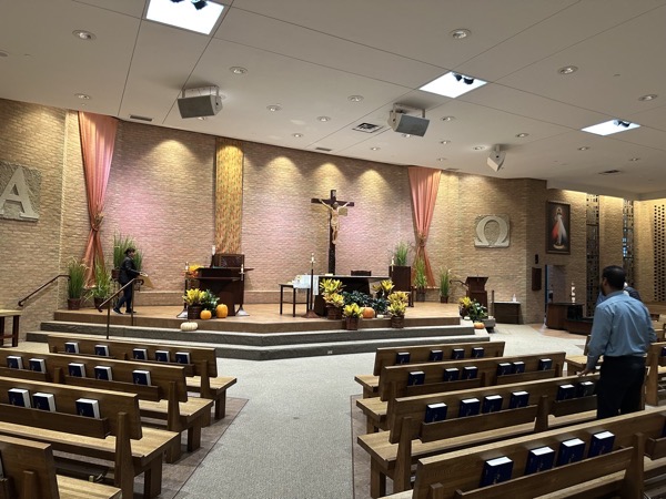 The interior of the church showing the sanctuary, a raised platform in the midst of the semi-circular post-Vatican II standard for a church. The back wall is brick with some curtains and stone bas relief carvings of alpha and omega along with a large crucifix. Most of the decoration in the church is two-dimensional, but there is a nook with a statue of the Holy Family. Also, atypical for this design of a church, there is a choir loft in the back of the church.