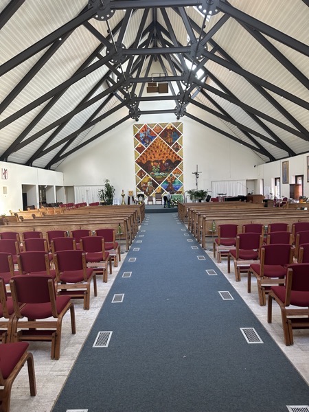 The interior of the church. A tapestry with a number of scenes of Catholic imagery in a African folk–inspired style covers the wall behind the sanctuary. The figure of Jesus on the Crucifix is semi-abstract, something only appaent on closer examination and the curved metal motif is repeated in the ambo.