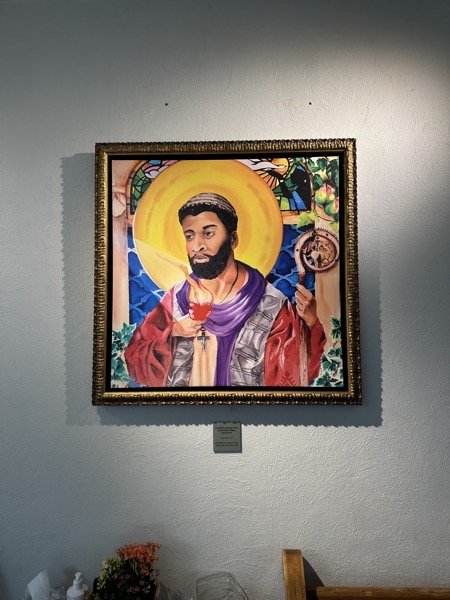 A hanging painting in the vestibule of the church, depicting St Augustine with African features. The label below it reads: An Unlikely Aquilegia: North Africa Saint of Hipopo, Canvas Print. Vernon Adams Artist, commissioned by Villanova University