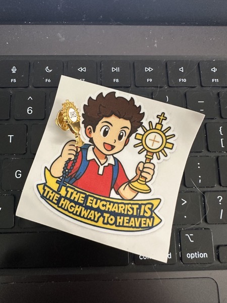 A sticker with a cartoon image of Carlo Acutis  with the message, “The eucharist is the highway to heaven” and a lapel pin of a monstrance sitting on the keyboard of my computer—I figured since Carlo Acutis is the patron saint of programmers and the internet, my computer keyboard would be a good backdrop for this photo.