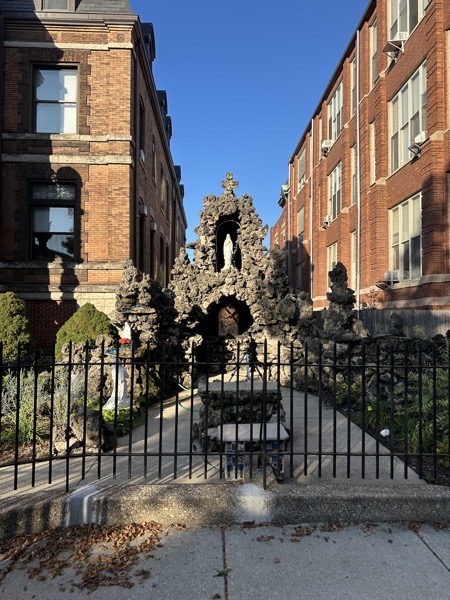 A grotto with a statue of the Virgin Mary in an upper alcove below a stone cross and a bas relief sculpture of Jesus carrying his cross in the small cave below the alcove. A stone prie-dieu stands before the grotto. In the background the rectory is on the left and an apartment building is on the right but the overall effect is as if the grotto were built in an alleyway between two apartment buildings.