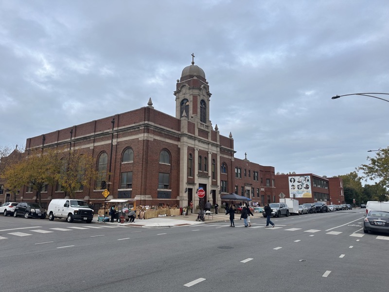The exterior of the church. A vendor of religious goods is set up on the sidewalk next to the church and anotehr vendor selling flavored waters is across from that vendor. The building itself is made of red brick and granite with a domed bell tower over the main entrance,