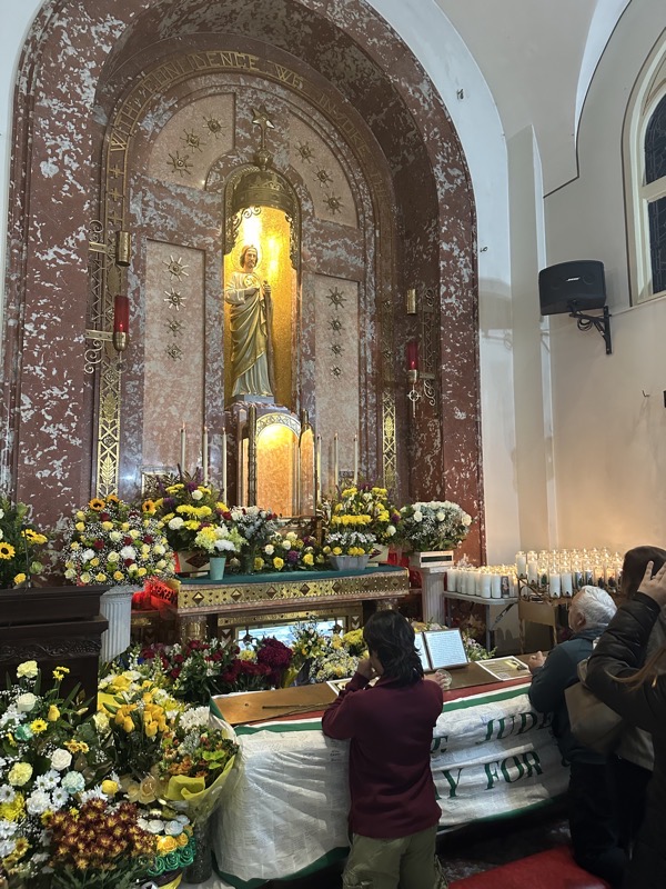 The side altar to St Jude with numerous flowers before it. The glass case below the statue of St Jude normally holds a reliquary with a piece of bone attributed to the saint, but it was removed for the procession through the nighborhood streets that took place after the Mass.