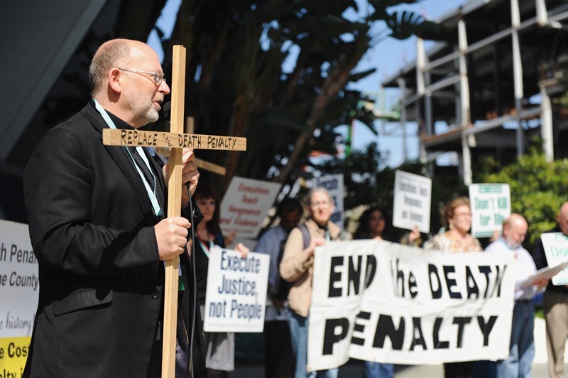 A photo of Father Chris speaking at an anti-death penalty protest in 2017. He is a bald white man with glasses and grey-brown hair wearing a black suit and holding a wooden cross on which is written “replace the death penalty”