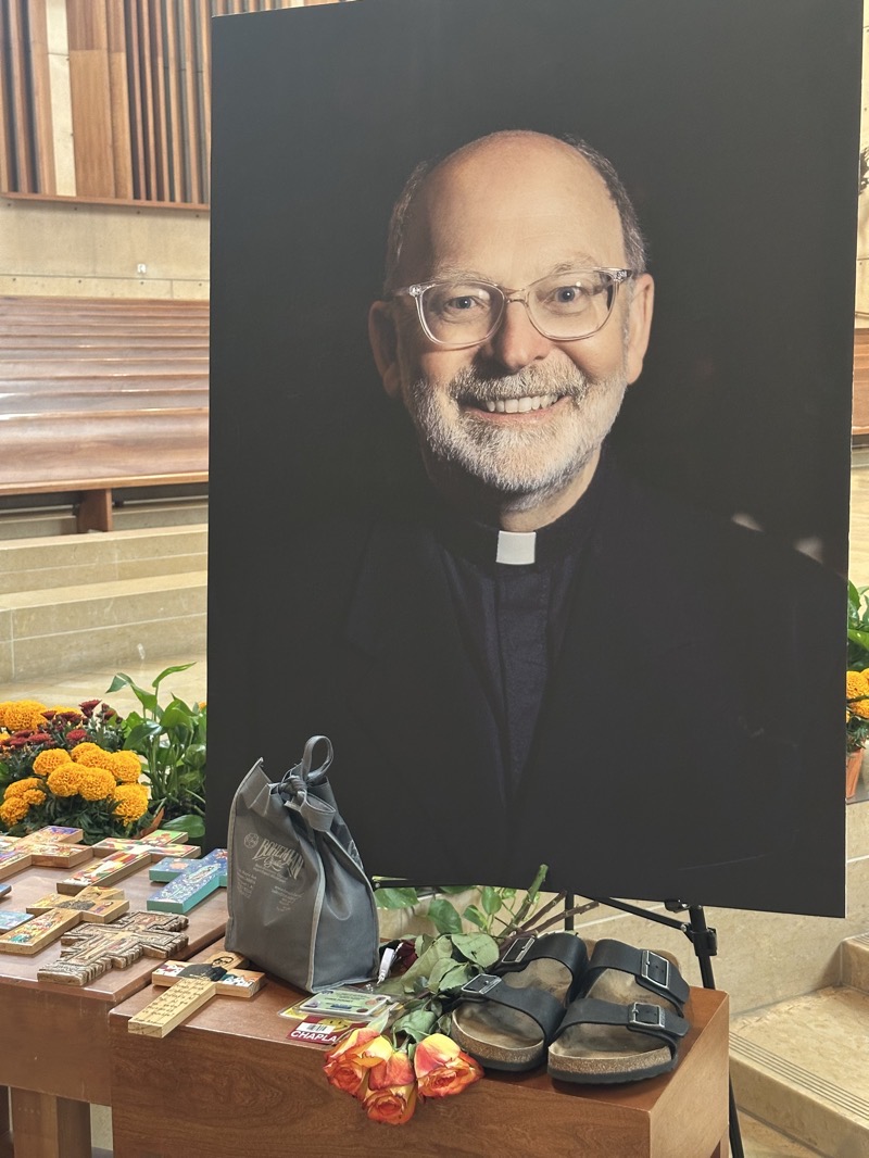 IMG_0961.jpeg Father Chris’s photograph with his assorted symbols in front of it. The photo show him smiling with glasses, a greying beard, bald head, black shirt with roman collar. Before him are a number of crosses painted in the San Damiano style, some flowers, his hospital badge, and crystal chalice in a black cloth bag.