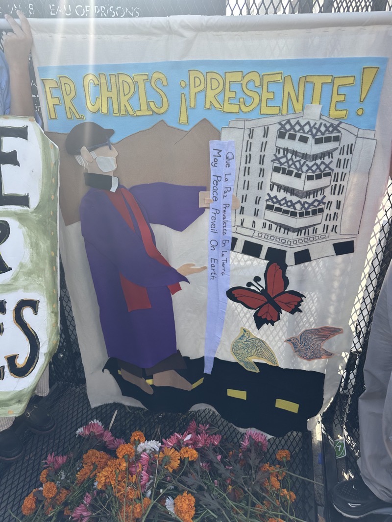 IMG_0965.jpeg a banner depicting Father Chris, standing before a peace pole with the metropolitan detention center in the background, a butterfly and two doves in the foreground. In front of the banner are flowers laid before the banner in memoriam of Chris at the conclusion of the protest/vigil.