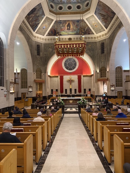 The interior of the shrine showing the sanctuary which is surrounded on four sides by pews, although the pews between the entrance and the sanctuary make the largest portion of the congregational seating. The church is decorated with mosaics in the domed ceiling and is in a sort of neo-medieval style.