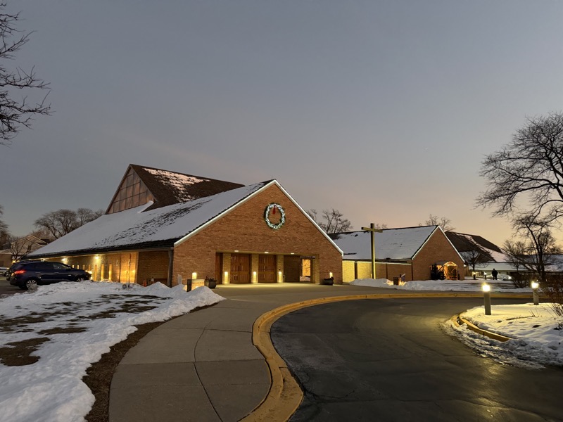 IMG_1099.jpeg The exterior of the church at night, a brick A-framed structure with a higher pitched roof which rises over the worship space