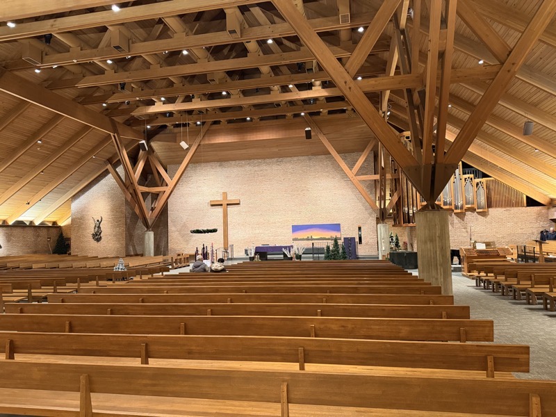 IMG_1100.jpeg The interior of the church which features exposed wooden beams. A plain wooden cross stands where a crucifix would be. On the wall behind the sanctuary there is a painting of a desert landscape under a nighttime sky. On the right side are figures representing the Holy Family, on the left are the figures of the three kings approaching the Holy Family. All the figures are separate objects attached to the picture and are moveable. Presumably, the Kings are moved across the picture until Epiphany and the baby Jesus will be added on Christmas.
