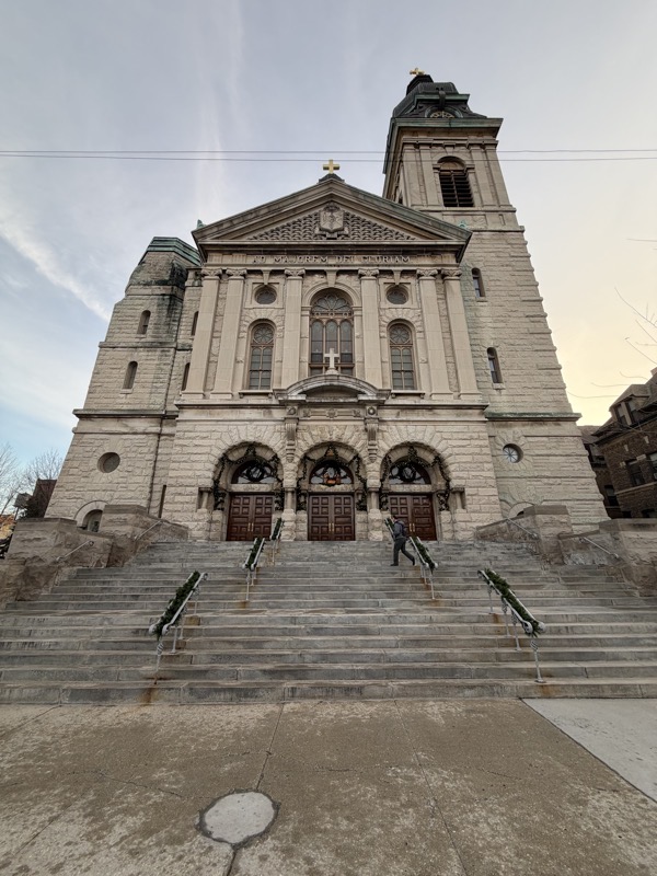 The front of Saint John Cantius Church, a neo-baroque building with a tall bell tower on the right side and the words “Ad Majorem Dei Gloriam” 