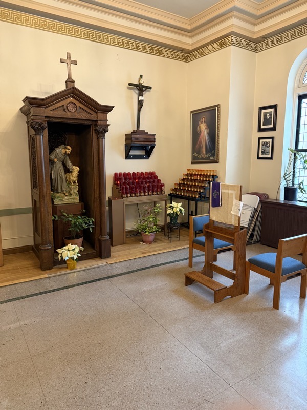 The confessional area at the back of the church. The original confessional has been repurposed to hold a statue of a saint (Francis?) while confessions are now held nearby in a pair of facing chairs or, for those who prefer, a kneeler with a screen separating the penitent from the priest
