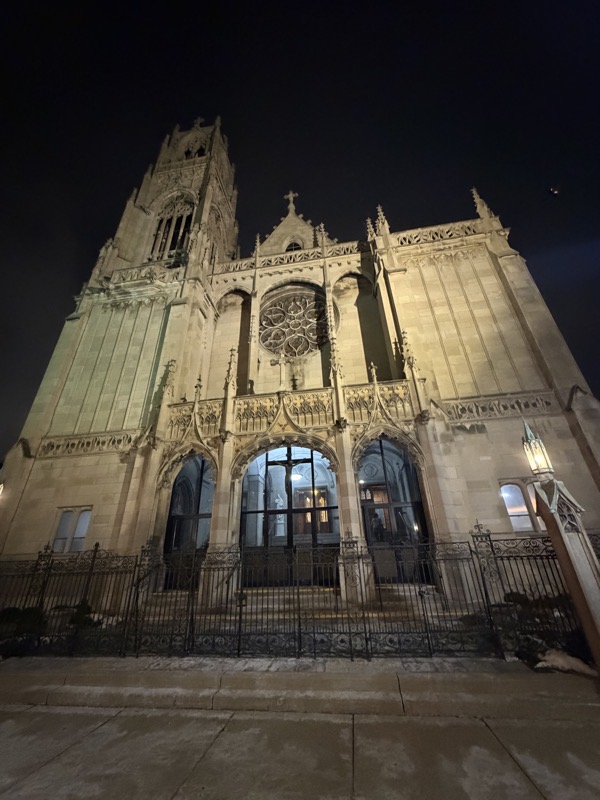 The exterior of St Ita lit dramatically in the night sky. It’s a neo-gothic style church with a bell tower on the left. The gates around the front entrance are padlocked shut and entry to the church at night is through a door on the side of the church