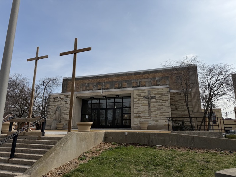 IMG_1464.jpeg Photograph of the exterior of the church, Stairs lead up to a rectangular brick building with a brick façade and a flat roof. A Crucifix is on the right of the glass entryway and a statue of St Richard to the left. A steel cross stands is positioned over the entrance. At the top of the stairs, two wooden crosses, erected for Lent, tower.