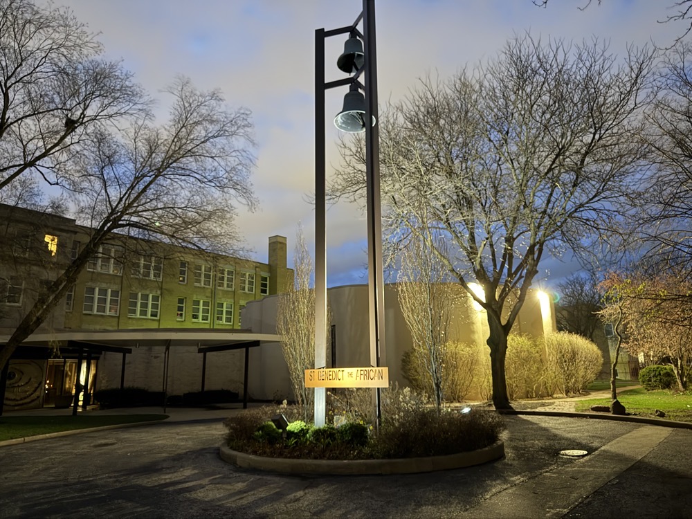 IMG_1469.jpeg The exterior of the church at night, showing the stand-alone belltower, older buildings of the church complex in the background and a round building in the foreground housing the church