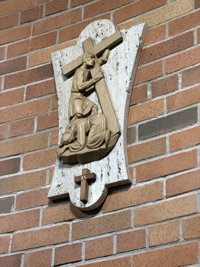 One of the stations of the cross. These are bas relief sculptures in wood. This one depicts Jesus falling for the third time, with another condemned prisoner carrying his cross while Jesus lies on the ground before him.
