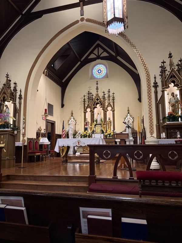 The view of the sanctuary from a pew near the front of the church. An altar rail separates the sanctuary from the nave. A pointed arch rises above the sanctuary. In the background, American and church flags can be seen.