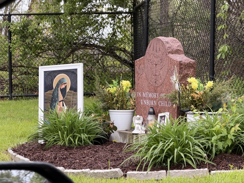 The tombstone dedicated to the memory of an unborn child surrounded by flowers, a picture of the Virgin Mary a couple small angel statues, another picture of Jesus, and I think a glass lantern with a candle inside, at least that’s what I hope it is. 
