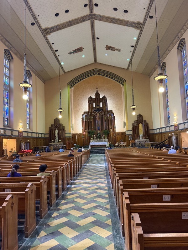 IMG_1606.jpeg The view of the sanctuary from the back of the church. The interior is relentlessly rectangular with a vaulted ceiling raising to a peak with angels painted on the ceiling panels. The old altar stands against the back wall of the sanctuary, with a large crucifix and statues of the four evangelists