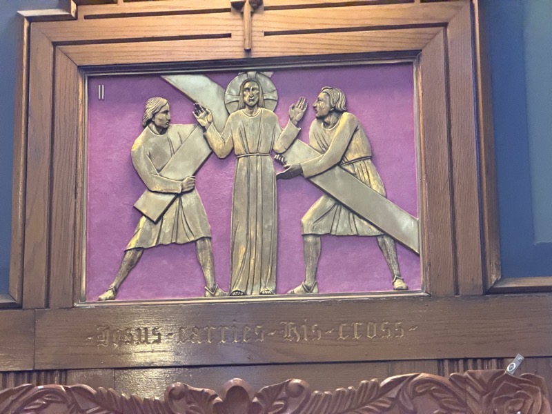 IMG_1608.jpeg One of the stations of the cross, a bas-relief engraving pained in gold against a light purple background. The station is “Jesus takes up his cross” but Jesus is standing before the cross with his arms raised while two anonymous figures actually lift the cross. It seems Jesus should be doing a bit more of the work here, I think.