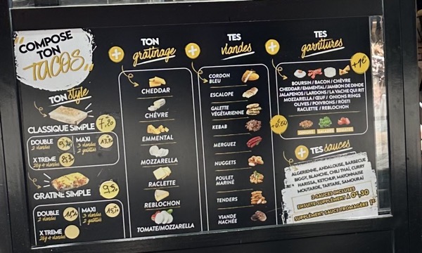 A menu board at a restaurant in Paris calling itself Chamas Tacos It offers a food item which appears to be a rectangular chunk of meet and cheese with optional add-ons including onion rings and sauces which include ketchup and mayonnaise all wrapped in what might be a wheat tortilla or might be a crepe. This is not a taco.